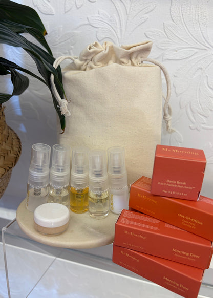 A skincare gift set featuring six clear sample spray bottles with yellow and white liquids, a small white jar, a cream canvas drawstring bag, and four orange boxes of Mr. Morning skincare, arranged on a clear acrylic stand.